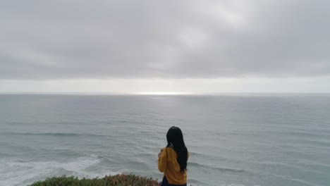 Reveal-of-Woman-Standing-at-Coastal-Cliff-on-the-Pacific-Taking-Photos-with-Phone-Reverse-Dolly