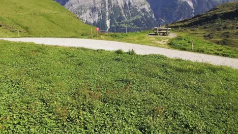 Aerial-Tilt-up-reveal-shot-of-a-hiker-sitting-on-a-Mountain-bench-by-a-hiking-trail-with-view-to-Snow-capped-swiss-alp-mountains-Schreckhorn-and-Finsteraarhorn-in-Summer
