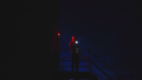 Mysterious-hooded-man-on-top-of-metal-stairs-shining-a-flashlight-around-at-night-time
