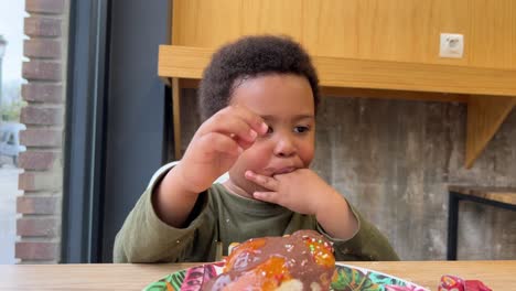 Funny-and-adorable-3-year-old-child-eating-a-cake-covered-with-chocolate-in-a-cafeteria-seated-next-to-his-mother