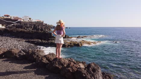 Caucasian-girl-in-summer-clothes-standing-on-beach-rocks-and-looking-at-ocean-waves