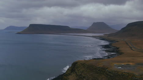 Waves-crashing-to-shore-below-a-mountain-with-a-roadway-curving-around-the-shore-and-several-vehicles-parked-at-a-lookout-point-in-Iceland