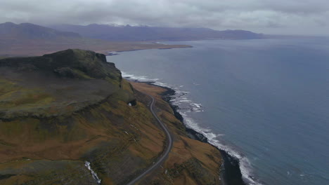 Vista-Pintoresca-De-Las-Olas-Rompiendo-En-La-Costa-Debajo-De-Una-Montaña-Con-Una-Carretera-Que-Se-Curva-Alrededor-De-La-Costa-En-Islandia
