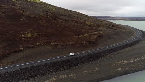 Aerial-Shot-of-Car-driving-between-a-Mountain-and-a-Lake