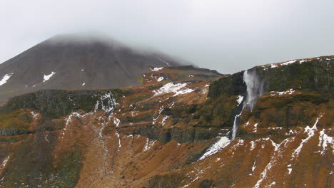 Malerische-Aussicht-Auf-Die-Berge-Mit-Atemberaubendem-Wasserfall,-Der-Von-Der-Spitze-Des-Berges-Herabstürzt-Und-In-Island-Nebel-Im-Wind-Weht