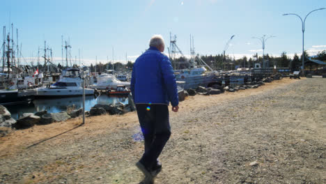 An-elderly-man-is-walking-on-a-pathway-in-Port-Alberni,-in-BC-Canada-and-countless-boats-and-blue-sky-in-the-background