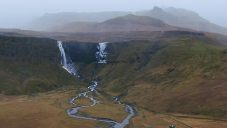 Drone-view-of-barren-ground,-mountains,-valleys-and-waterfalls-amidst-overcast-skies-in-Iceland-Kirkjufell-Mountain-near-Grundarfjordour
