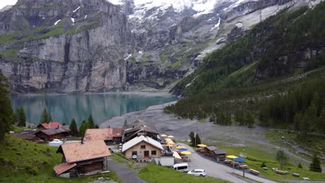 Antena-De-Drones:-Joven-Excursionista-Sentado-En-Una-Roca-En-La-Cafetería-De-La-Montaña-Frente-A-Las-Montañas-Gigantes-Bluemlisalp-Que-Rodean-El-Lago-Glaciar-Turquesa-Szure-Oeschinensee-En-Kandersteg,-Suiza