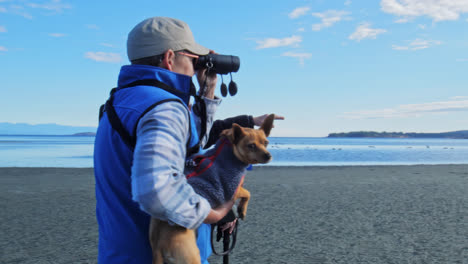 an-elderly-man-and-lady-look-in-the-distance-through-a-telescope-on-a-beach---A-man-holding-a-dog-looking-through-a-telescope-in-a-sunny-day