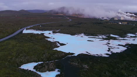 Road-curving-past-a-blue-lagoon-with-factories-and-smoke-in-the-distance-in-Iceland