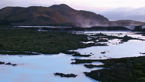 Impresionante-Vista-De-Una-Laguna-Azul-Con-Reflejos-Del-Cielo,-Vapor-Y-Montañas-En-El-Fondo-En-Islandia