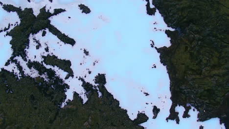 Aerial-top-down-view-spin-2-of-a-blue-lagoon,-snow-and-ice-covered-surrounded-by-rocky-terrain-in-Iceland