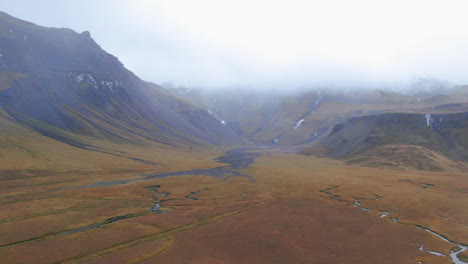 Slowly-zooming-in-drone-view-of-overcast-skies,-snow-kissed-mountains,-barren-ground,-valleys-and-streams-in-Iceland-Kirkjufell-Mountain-near-Grundarfjordour