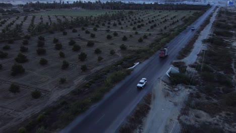 Aerial-drone-backward-moving-shot-over-cars-and-lorries-driving-along-RCD-road-with-farmland-on-both-sides-of-Baluchistan-during-evening-time
