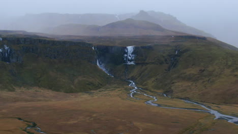 Aerial-view-zooming-in-of-overcast-skies,-mountains,-barren-ground,-valleys,-waterfalls-and-streams-in-Iceland-Kirkjufell-Mountain-near-Grundarfjordour