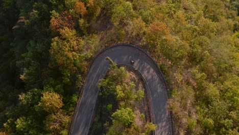 Aerial-top-down-view-of-motorist-making-a-turn-on-a-wooded-mountain-road-at-sunset