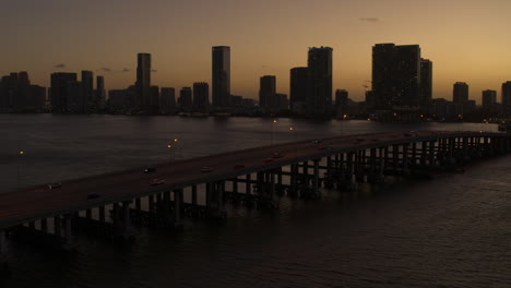 Miami-Bridge-Mit-Der-Innenstadt-Von-Miami-Im-Hintergrund-Und-Vorbeifahrenden-Autos-Bei-Sonnenuntergang,-Luftaufnahme