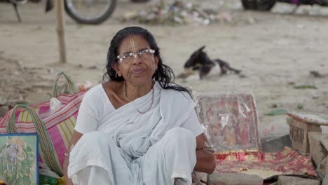 Devotee-Indian-old-woman-sitting-outside-near-temple-with-photo-frames-of-Gods,-slow-motion