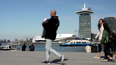 People-walk-in-foreground-of-GVB-ferries-in-water-by-Amsterdam-skyline