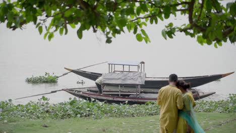 Indian-couple-in-traditional-wear-standing-near-river-bank-of-Ganga,-Rani-Rashmoni-ghat,-boats-in-background,-slow-motion