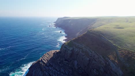 Panorama-Luftaufnahme-Einer-Dunstigen,-Sonnigen-Küstenlandschaft-In-Spanien,-Playa-De-Tagle,-Grüne-Wiesenhochebenen-Bilden-Felsige-Klippen,-Meereswellen-Des-Ozeans-Spülen-An-Die-Küste