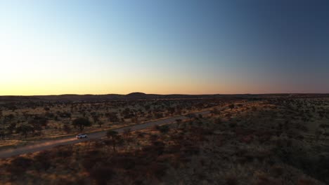 Drone-shot-following-a-white-SUV-moving-across-distant-prairie,-sunset-in-Namibia,-Africa