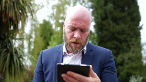 An-unhappy-man-in-a-business-suit-working-outside-in-a-garden-using-a-tablet-computer