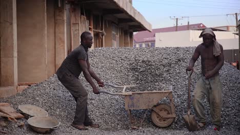 Construction-Worker-Using-Shovel-to-Fill-a-Wheelbarrow-with-stone-chippings-and-young-adult-pushes-wheelbarrow-away