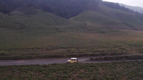 Drone-flies-around-yellow-jeep-thats-drives-on-dirt-road-at-mount-bromo-indonesia,-aerial