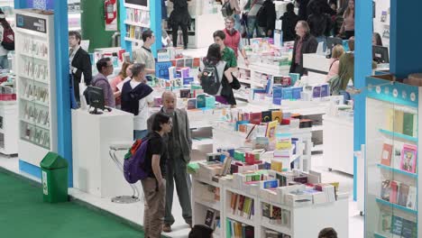 Estanterías-De-Libros-Brillantemente-Iluminadas-En-El-Centro-De-La-Feria-Del-Libro,-La-Gente-Camina-Buscando