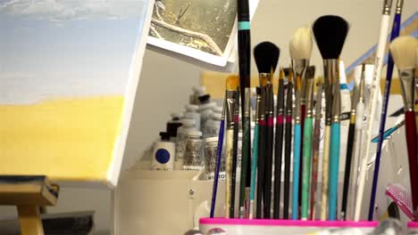 Pan-across-paintbrushes-stacked-on-artists-desk-towards-monument-valley-desertscape-painting