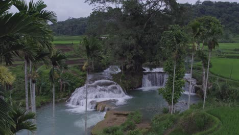 Sideways-shot-of-the-Waikelo-Sawah-Waterfall-with-palm-trees-in-front-at-Sumba-island-Indonesia,-aerial
