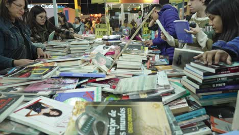 People-crowd-around-messy-stacks-of-books-on-table-flipping-and-searching-through