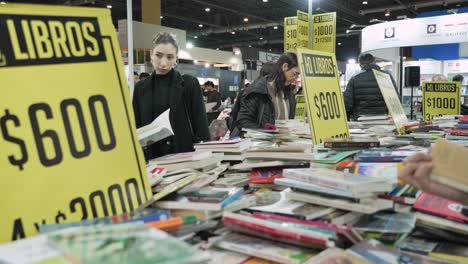 Zwei-Frauen,-Eingerahmt-Zwischen-Rabatttischschildern-Auf-Der-Buchmesse,-Mit-Willkürlich-Gestapelten-Büchern-Auf-Dem-Tisch