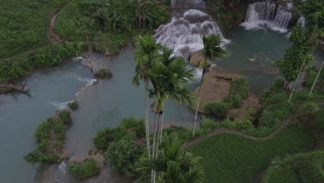 Drone-flies-close-to-palm-trees-next-to-Waikelo-Sawah-Waterfall-at-Sumba-Indonesia,-aerial