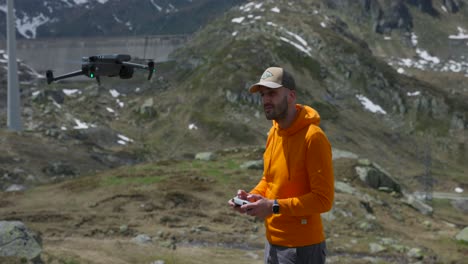 Drone-pilot-operator-in-orange-hoodie-and-cap-flies-drone-above-him-with-mountain-scenery-in-background