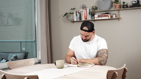 Hombre-Barbudo-Y-Tatuado-Escribiendo-Un-Diario-En-La-Mesa-En-Casa