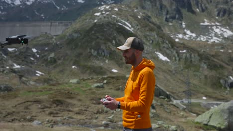 Profile-view-of-drone-pilot-operator-in-orange-hoodie-and-cap-flying-drone-above-him-with-mountain-scenery-in-background