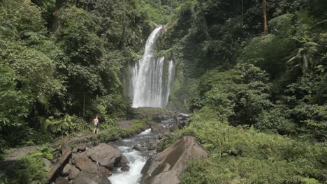 Luftaufnahme:-Männlicher-Tourist-Joggt-Am-Fluss-Entlang-Zum-Fuß-Des-Wasserfalls,-Lombok