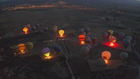 night-scene,-drone-camera-is-flying-over-a-valley-in-Cappadocia,-Turkey,-with-a-number-of-hot-air-balloons-flashing-lights,-causing-the-balloons-to-rise,-with-a-city-view-in-the-background-and-sunset