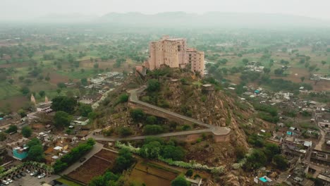 established-shot-aerial-drone-camera-moving-back-showing-Leela-Bisangarh-Jaipur-fort-with-only-one-way-to-go-up-or-down-with-vehicles-passing-by-and-core-residential-houses-surrounding-the-fort