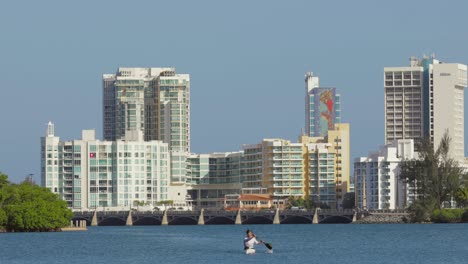 Person-kayaks-in-open-water-with-Hilton-hotels-and-Puente-Dos-Hermanos-of-the-Condado-area-of-the-city-in-the-background---Pan-right