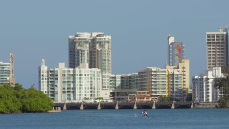 Person-kayaks-in-open-water-with-Hilton-hotels-and-Puente-Dos-Hermanos-of-the-Condado-area-of-the-city-in-the-background---Pan-right