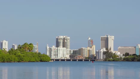 Person-kayaks-in-open-water-with-Hilton-hotels-and-Puente-Dos-Hermanos-of-the-Condado-area-of-the-city-in-the-background---Pan-right---Wide-Shot