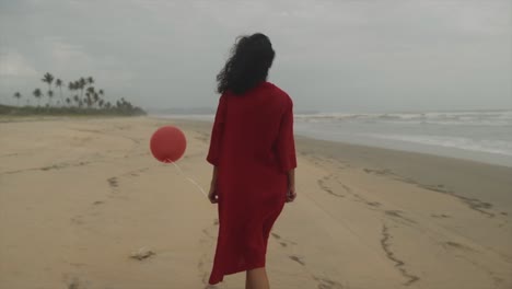 Beautiful-Young-Indian-brunette-Woman-in-a-red-dress-with-a-red-balloon-in-hand-walking-along-the-beach-of-the-Arabian-Sea-in-Goa,-India,-on-a-cloudy-day
