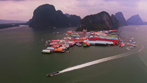 Panyee-floating-village-aerial-view-in-which-a-boat-full-of-men-is-going-to-the-sea-at-full-speed-and-behind-it-a-large-white-color-shed-is-falling-in-the-water-which-looks-landscape-and-beautiful