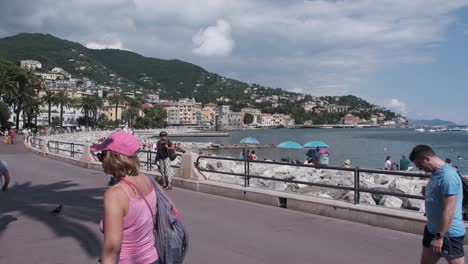 Gente-Caminando-Por-El-Paseo-Marítimo-De-Rapallo,-Italia,-Mientras-Otros-Toman-El-Sol-En-La-Playa.