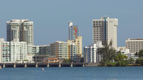 Person-kayaks-in-open-water-with-Hilton-hotels-and-Puente-Dos-Hermanos-of-the-Condado-area-of-the-city-in-the-background---Pan-right---Wide-Shot