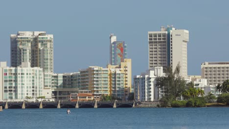 Persona-Kayaks-En-Aguas-Abiertas-Con-Los-Hoteles-Hilton-Y-El-Puente-Dos-Hermanos-Del-área-Del-Condado-De-La-Ciudad-Al-Fondo---Panorámica-Hacia-La-Derecha---Plano-Amplio