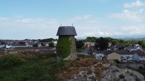 Aerial-view-passing-Melin-Wynt-Y-Craig-disused-Llangefni-windmill-ivy-covered-hillside-landmark-to-reveal-Welsh-Snowdonia-mountains,-Anglesey